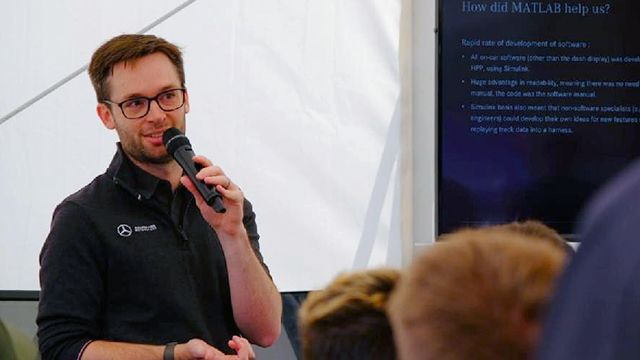 Graham, wearing glasses and a dark button-down shirt, stands in front of a seated audience and holds a microphone to his mouth.