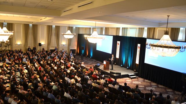 A large audience listens to a speaker presenting on stage at a professional conference in a hotel ballroom. 