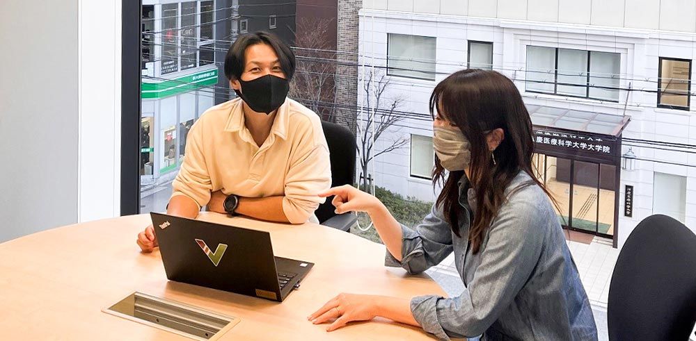 Two people wearing masks and sitting at conference table, working together on a laptop.