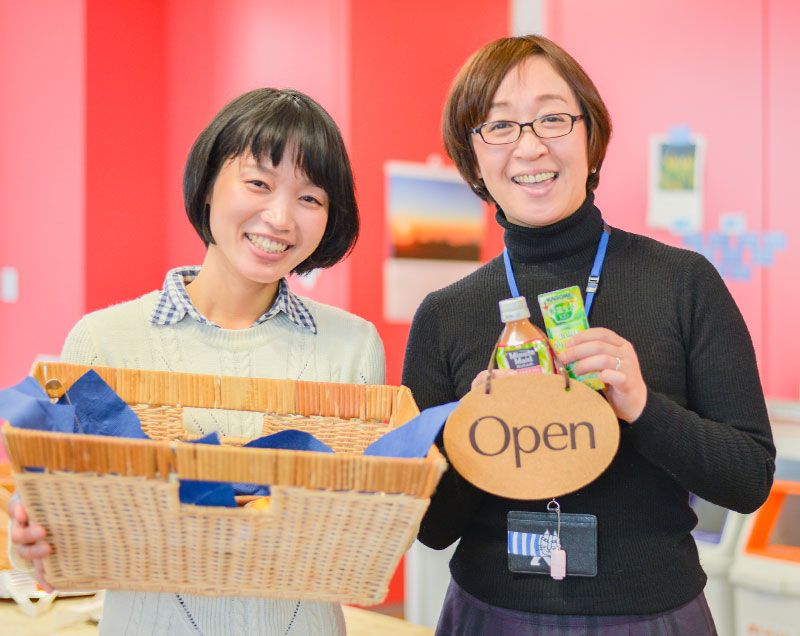 Two women, both smiling, one holding a basket of fresh baked goods and the other holding a bottled drink.