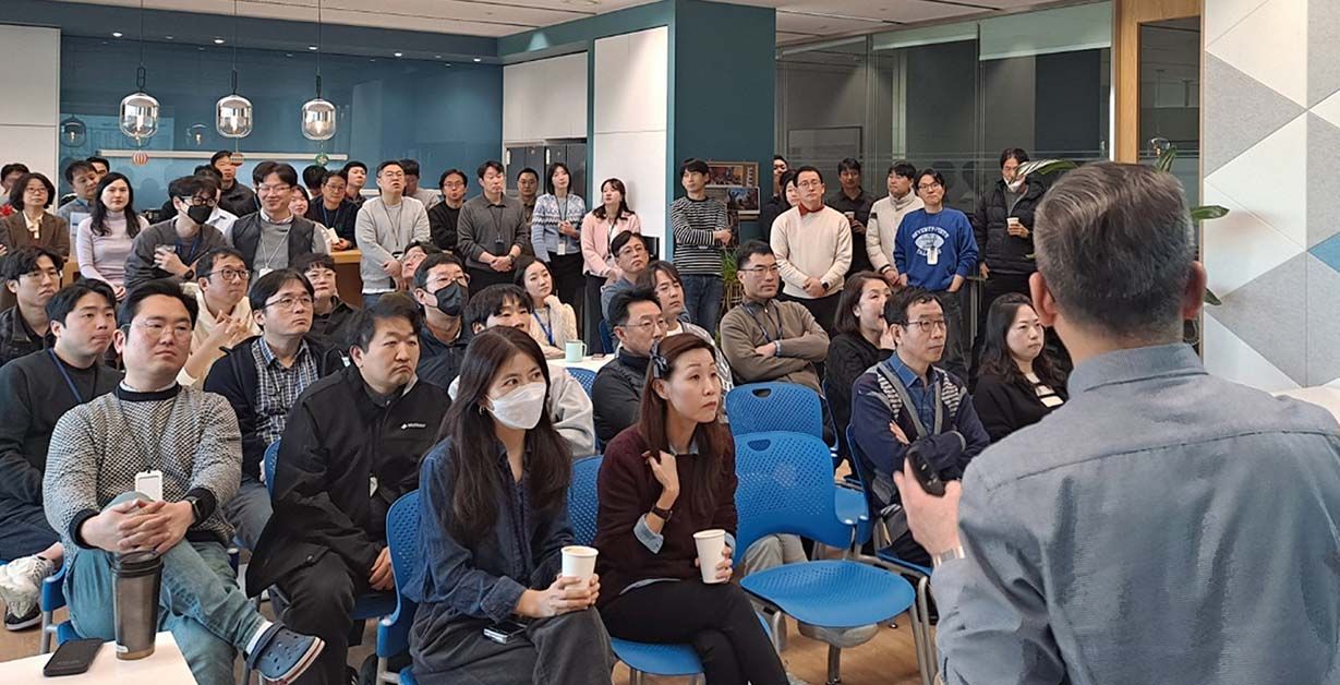 A large group of people, seated and standing, attentively listen to a speaker in a conference room.
