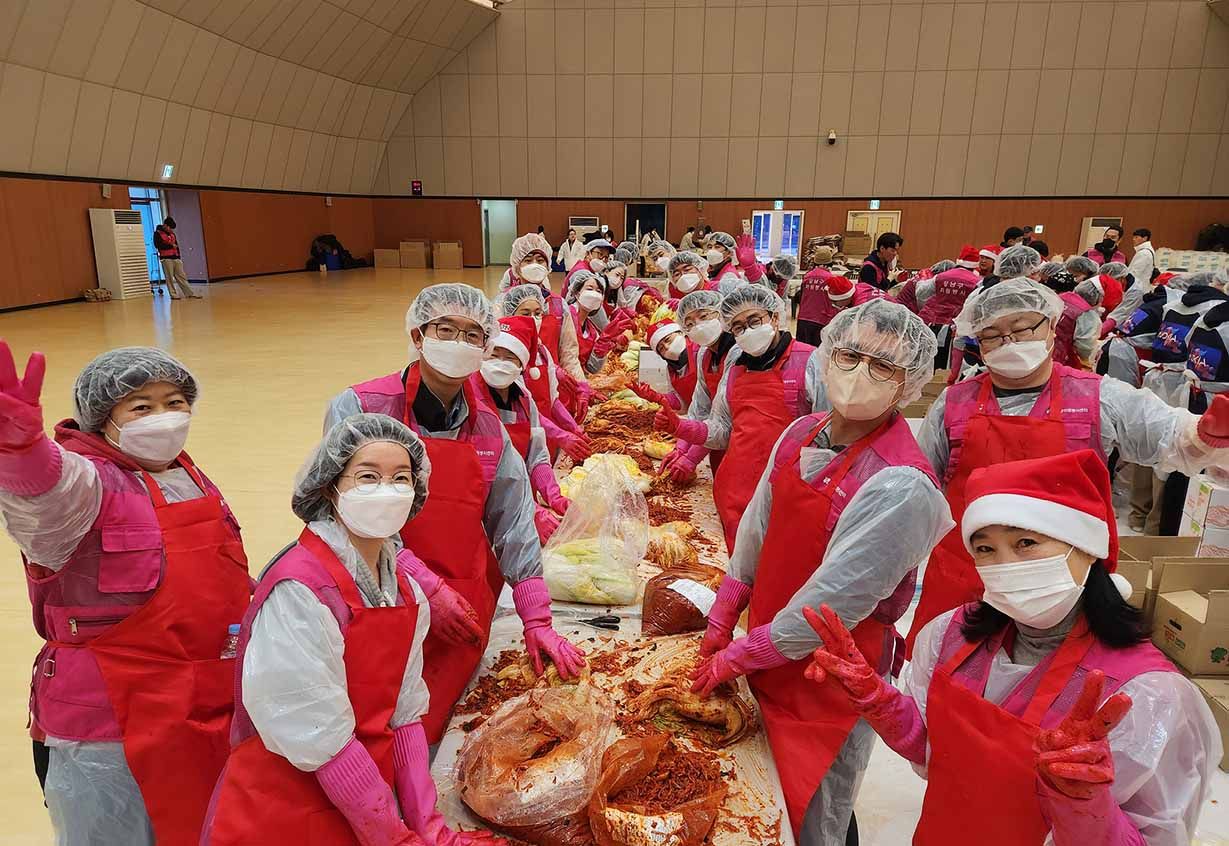 Volunteers in red aprons, gloves, and hairnets make kimchi together at a long table in a large indoor hall.
